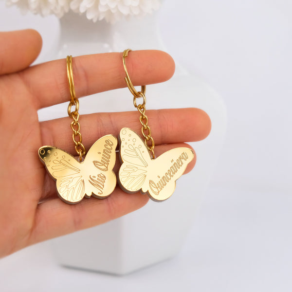Gold keychain butterfly-shaped charms with engraved names on a hand against a light background