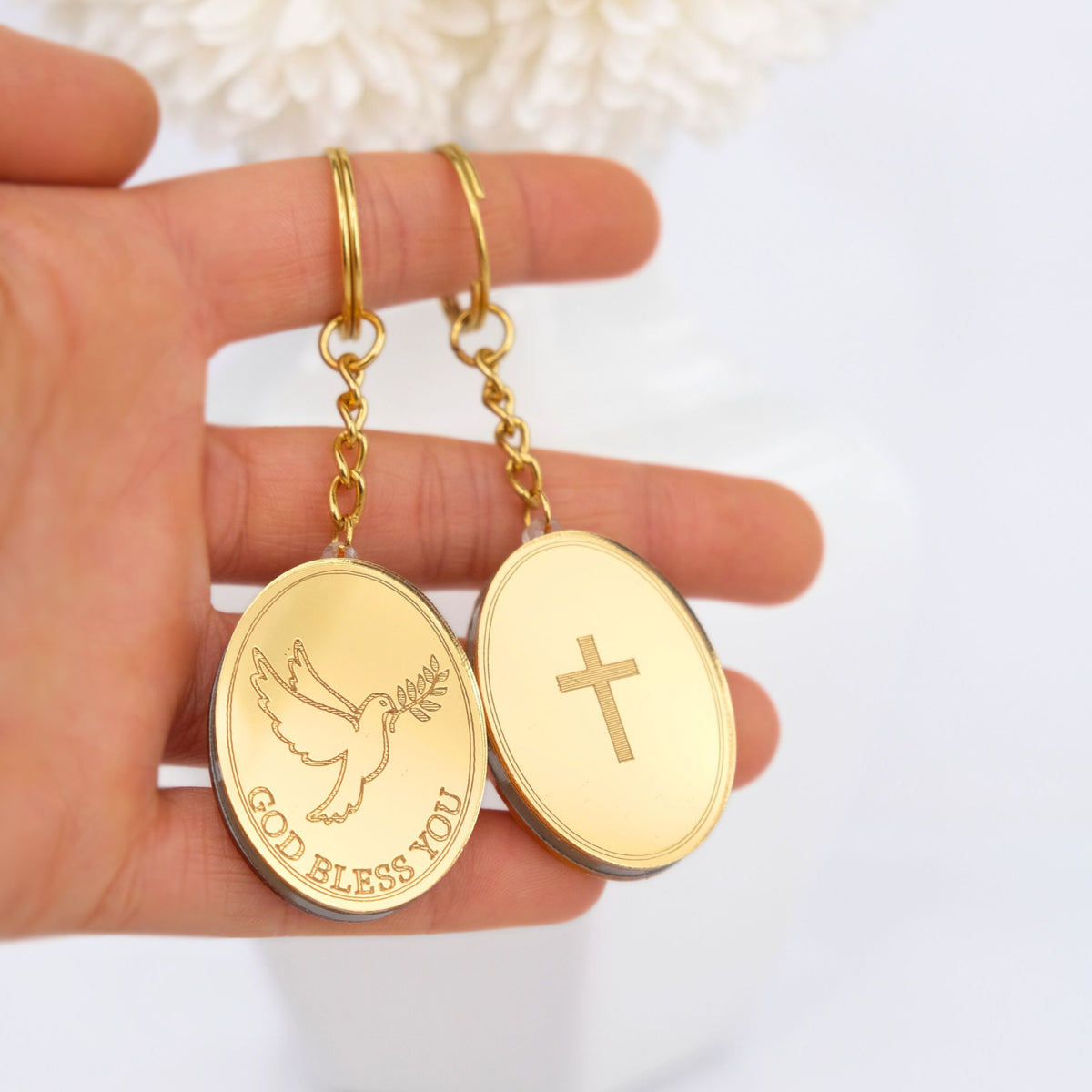 Gold keychain favor pendants with 'GOD BLESS YOU' and cross designs held in a hand against a white background.
