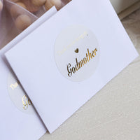 Sticker with 'Godmother' text on a white card, with wooden beads in the background.