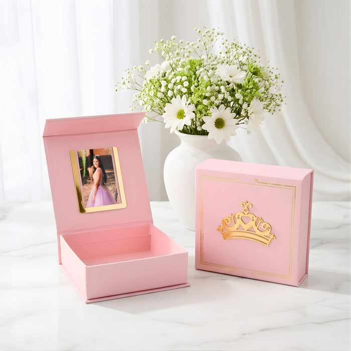 Pink gift box with a gold crown design next to a vase of flowers on a white surface.