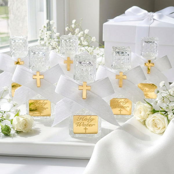 Holy water bottle favors with gold crosses and 'Holy Water' labels on a white surface with flowers.