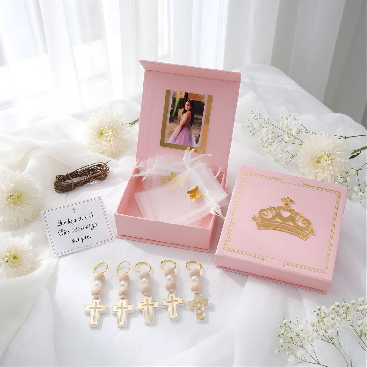 Pink gift box with gold cross charms and decorative quinceanera gifts on a white surface with flowers.