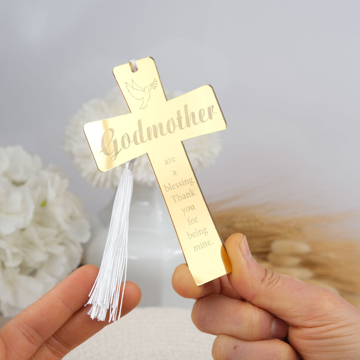 Gold cross with 'Godmother' engraving held by a hand against a blurred floral background