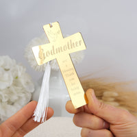 Gold cross with 'Godmother' engraving held by a hand against a blurred floral background