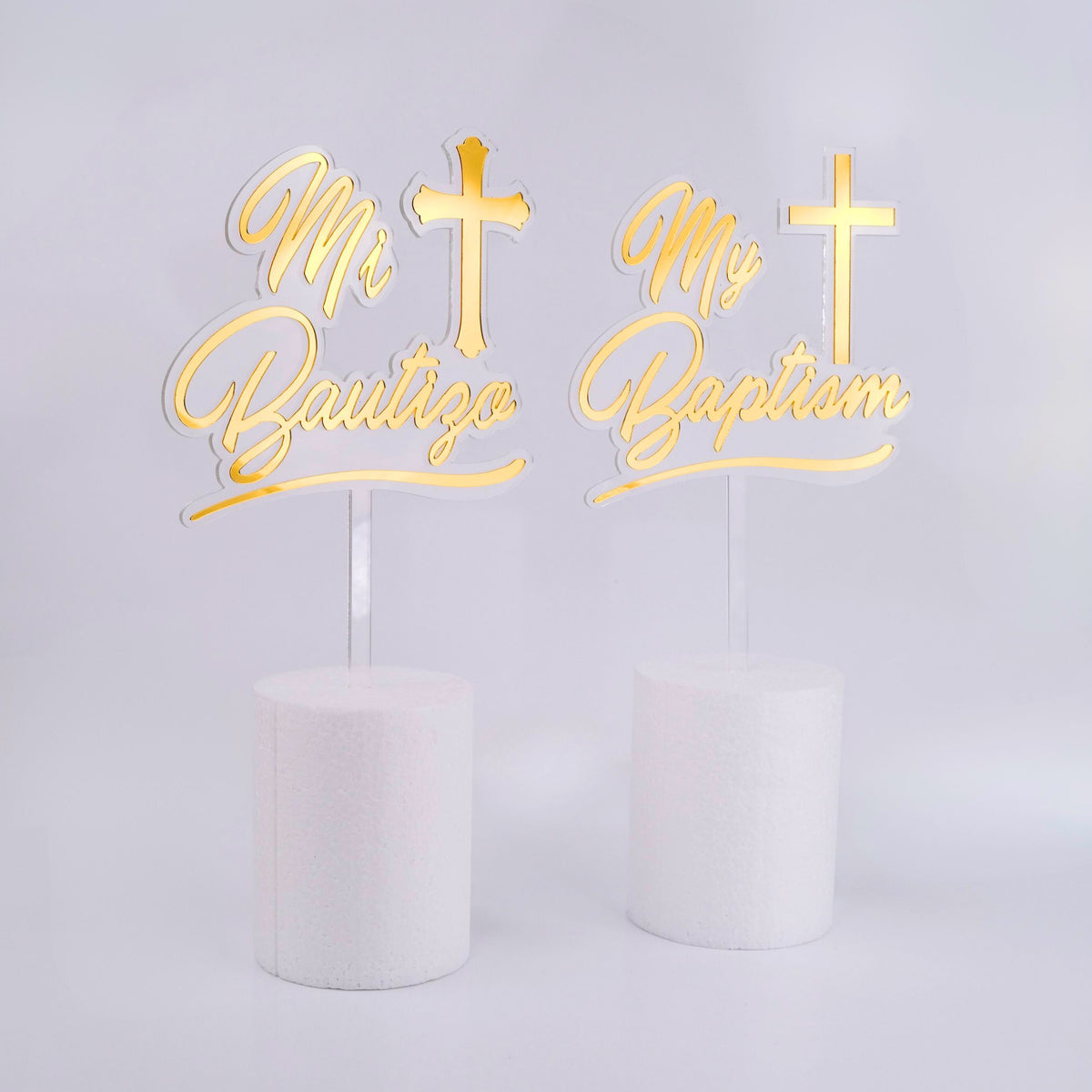 Two gold cake toppers with crosses and text on white cakes against a light gray background