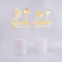 Two gold cake toppers with crosses and text on white cakes against a light gray background