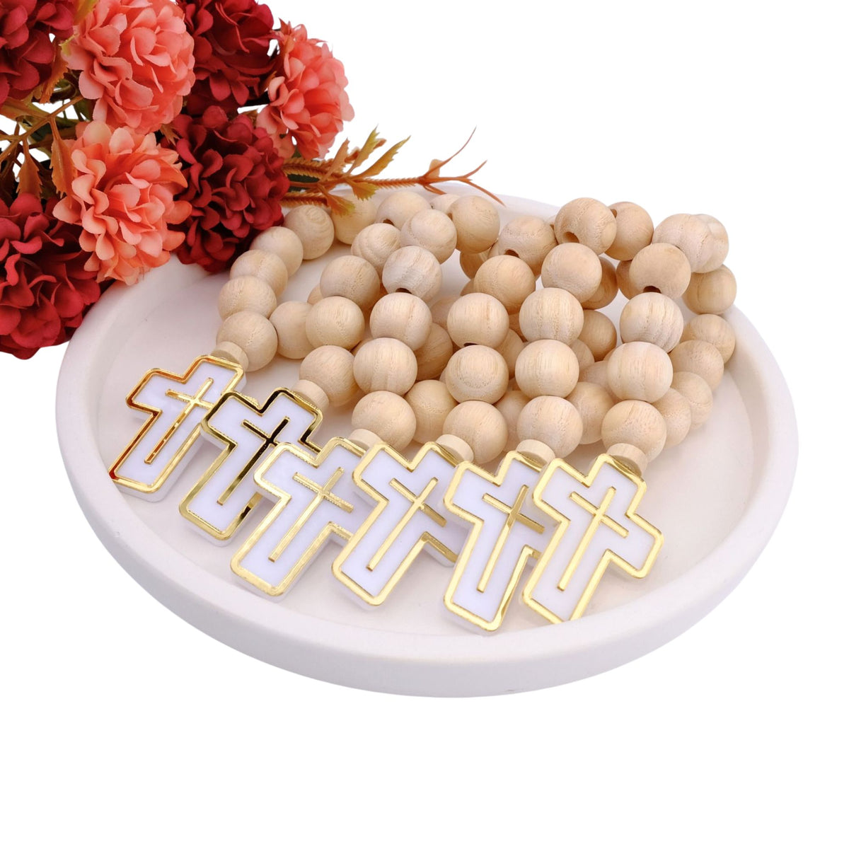 Wooden beaded mini rosary favors with white and gold lettered pendants on a white dish with red and pink flowers.