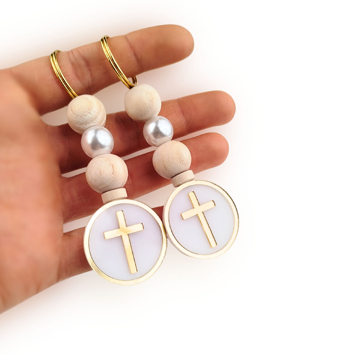 Hand holding a pair of keychain favors with wooden beads and gold crosses on a white background