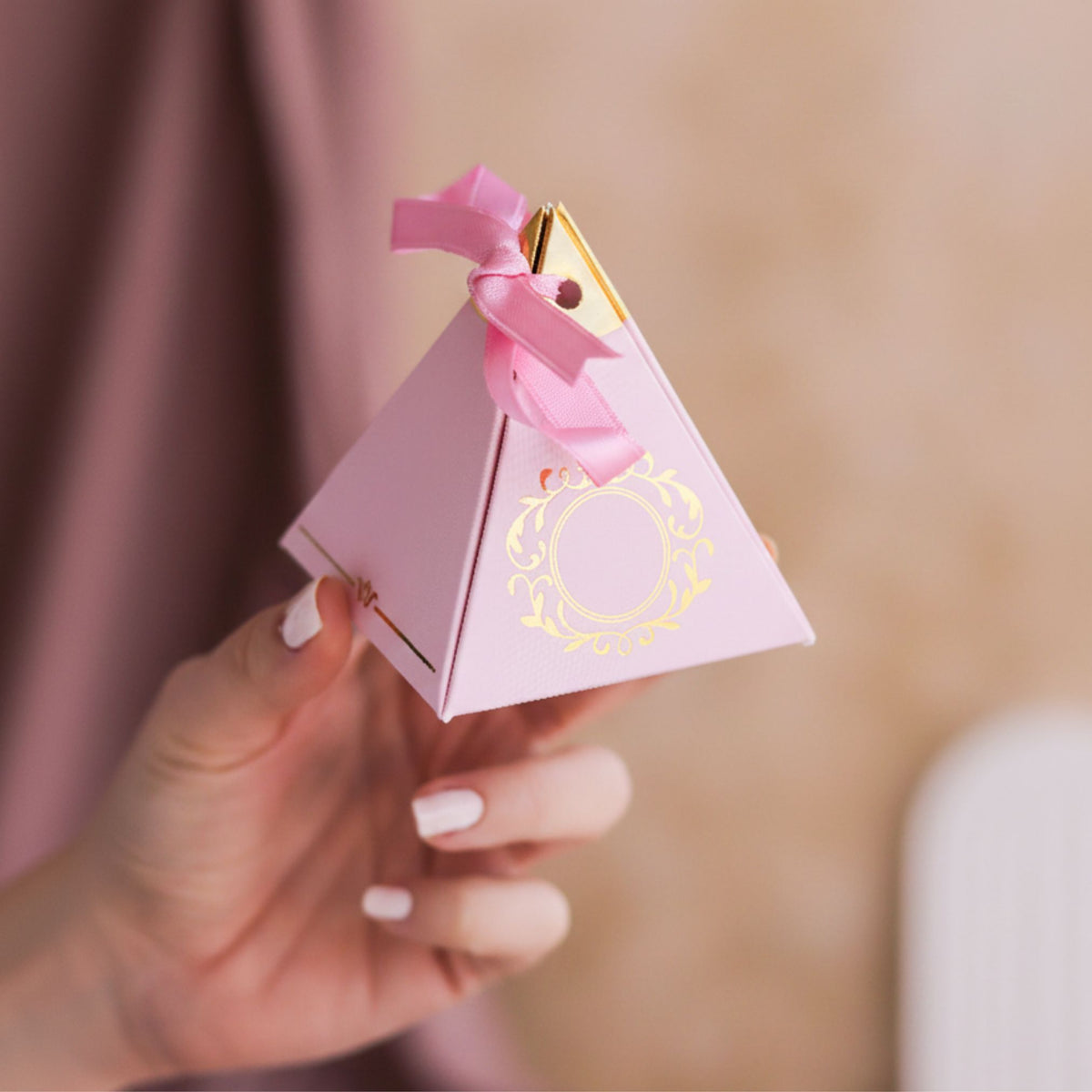 Pyramid-shaped pink gift box with gold accents and a pink ribbon held by a hand against a blurred background.
