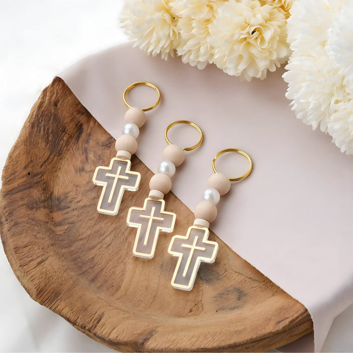 Three cross-shaped keychains with beads on a wooden surface with flowers in the background