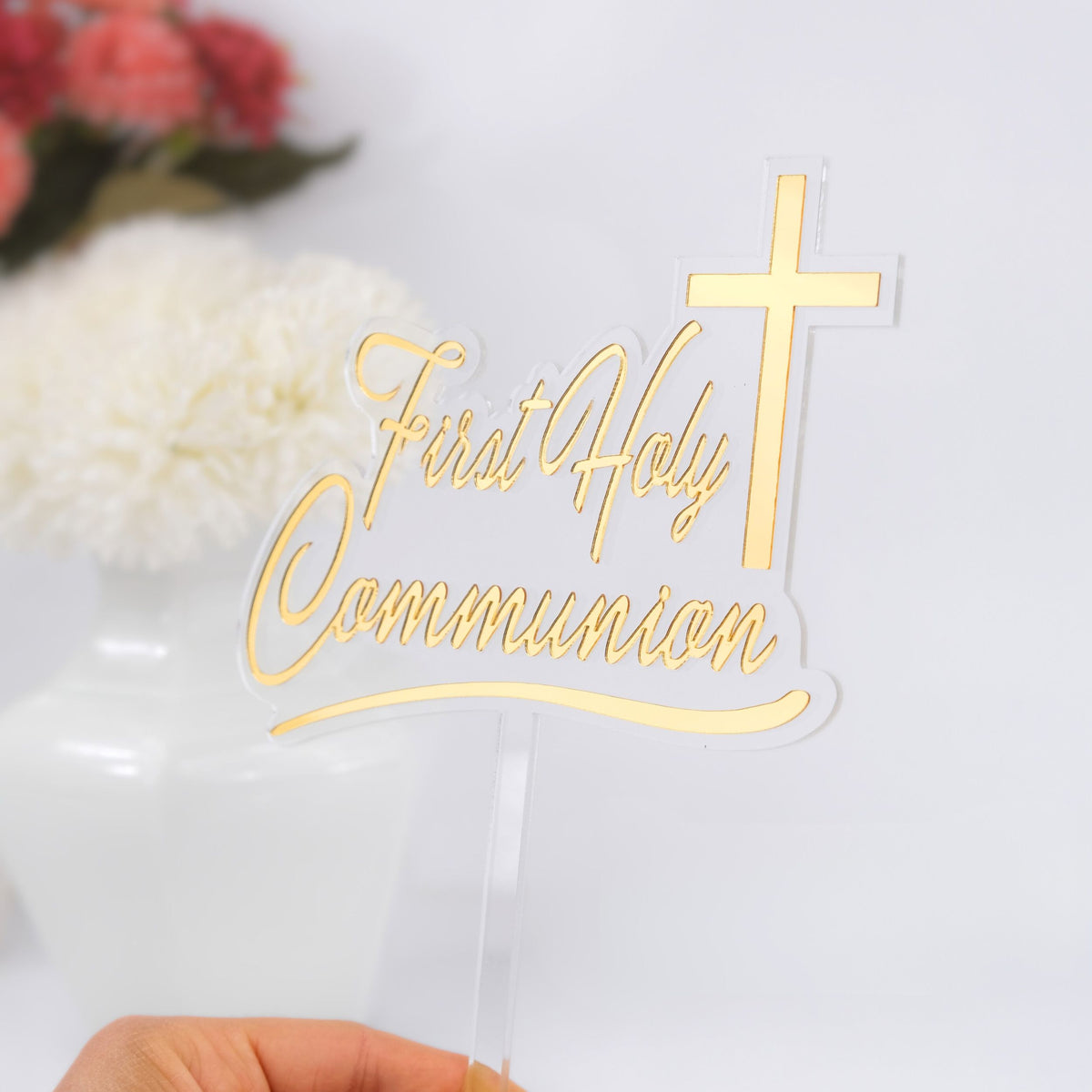 Clear cake topper with gold 'First Holy Communion' text and cross, held by a hand.