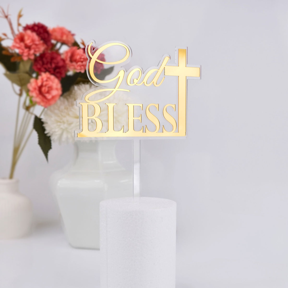 Gold 'God Bless' cake topper with a cross on a white cake against a light background with flowers.