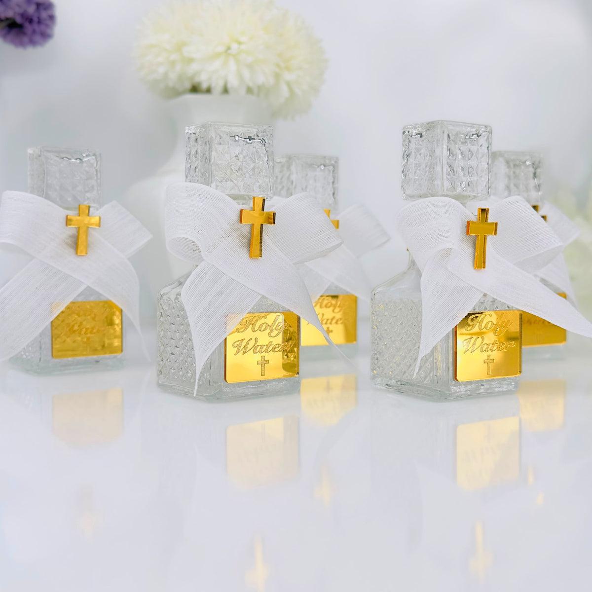 Three small glass holy water bottles with gold crosses and white ribbons on a reflective surface.
