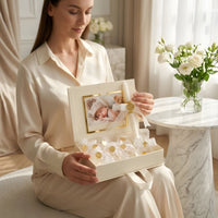 Woman holding a decorative baptism gift box with baby photos and keepsake items in a cozy room.