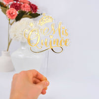 Gold 'Mis Quince' cake topper with a crown, held by a hand in front of a vase with flowers.