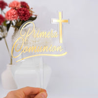 Gold 'Primera Comunion' cake topper with a cross, held in front of flowers.