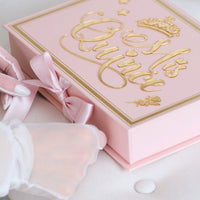 Pink gift box with gold text and a pink ribbon, held by a gloved hand.